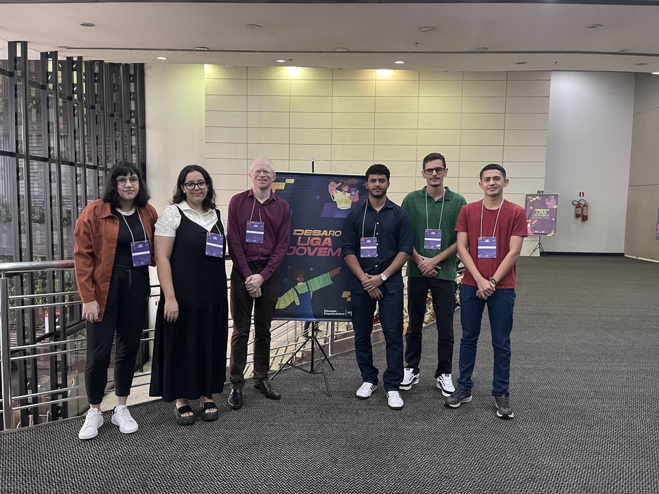 Foto colorida com os estudantes e professor orientador ao lado de uma bnner do evento.